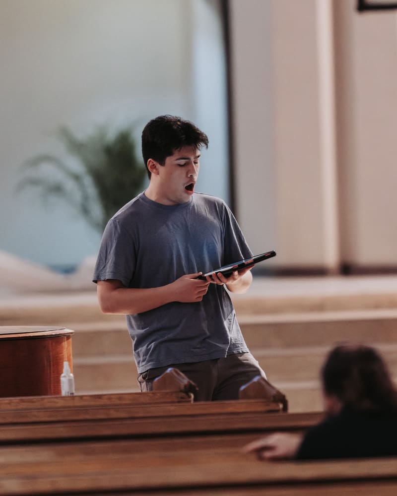 Matthew Menard singing from a folder, standing behind church pews